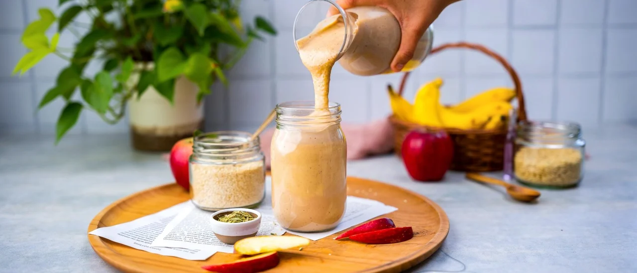 Sesame apple smoothie being poured into a glass jar, with apples, bananas and sesame seeds